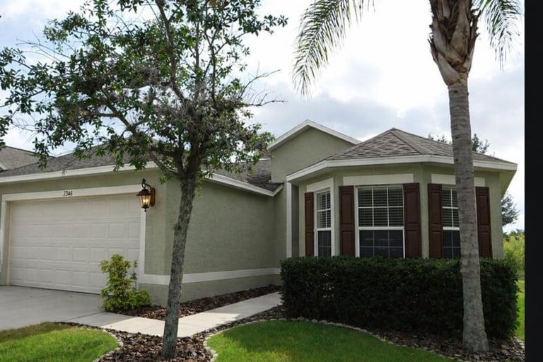 Single-story house with tan exterior, brown trim, attached garage, and palm trees in landscaped yard