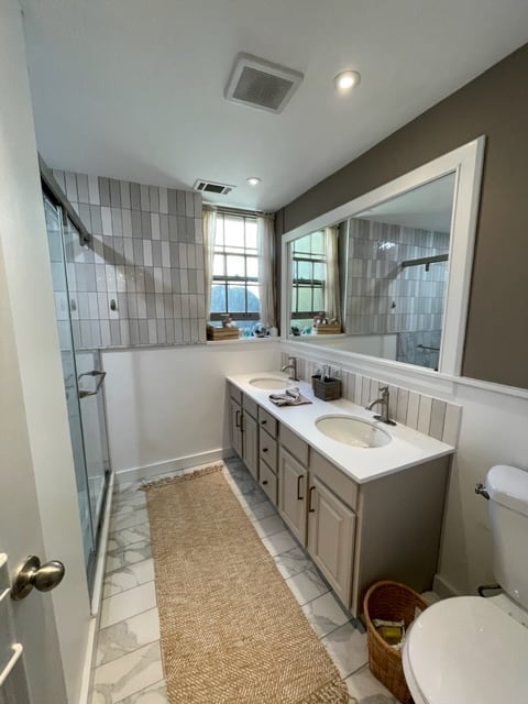Modern bathroom with gray and white tile walls, large mirror, white vanity with sink, toilet, and walk-in shower with glass door
