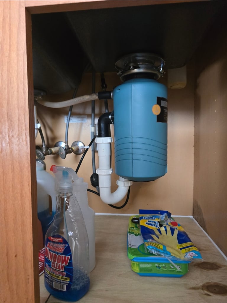 Under-sink garbage disposal with blue canister mounted on cabinet wall, pipes and household cleaning supplies stored below